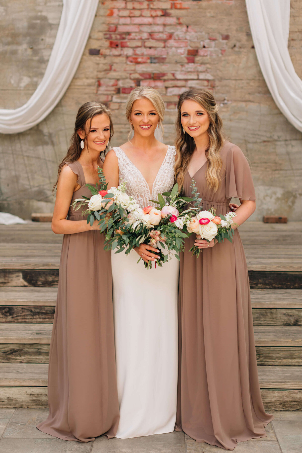 Three women in formal dresses stand together, smiling and holding bouquets. The woman in the center wears a white wedding dress, while the two on either side wear matching taupe dresses. They stand in front of wooden steps and a brick wall.