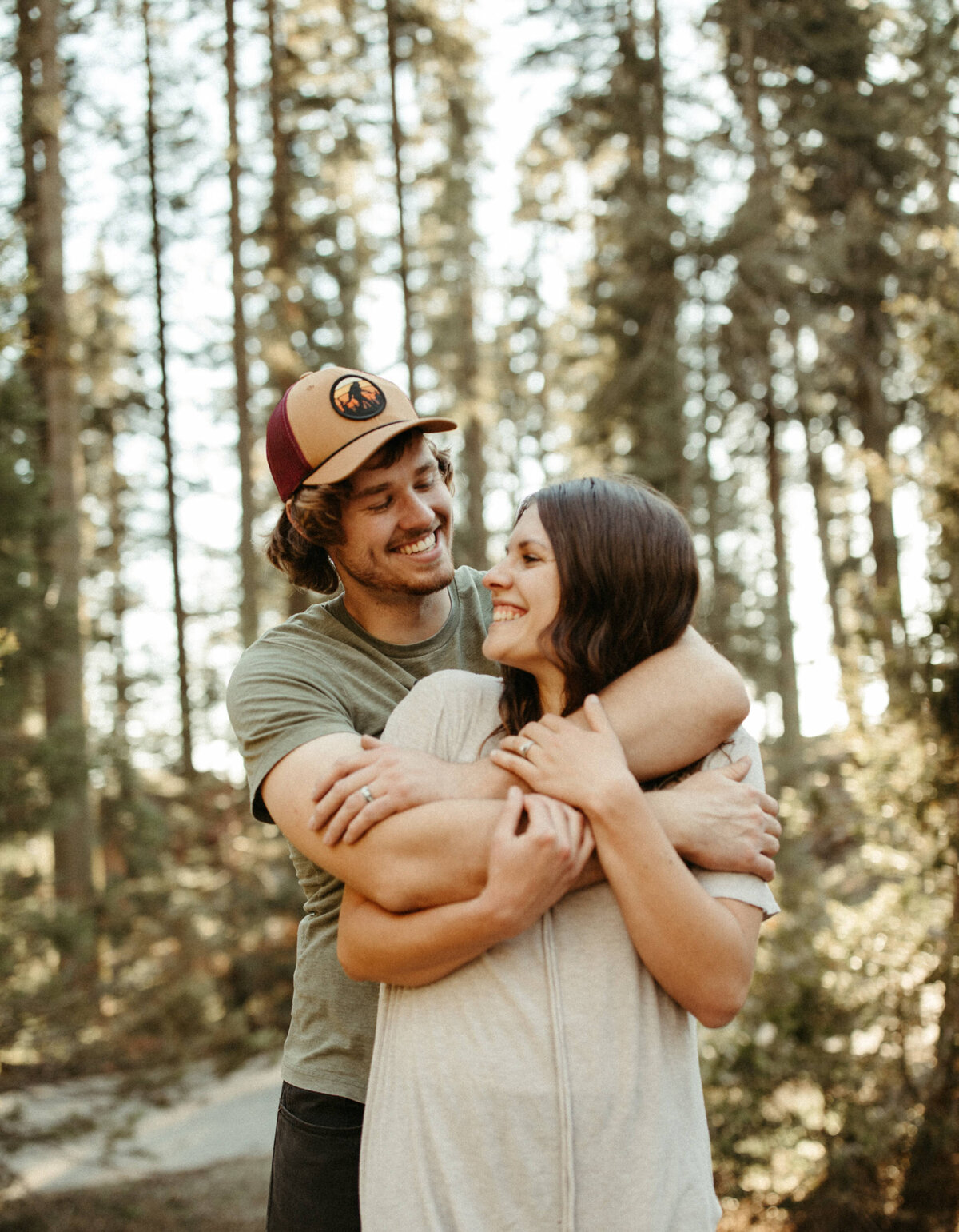 Guy in hat wrapping his arms around a girl in the woods with tall trees behind them