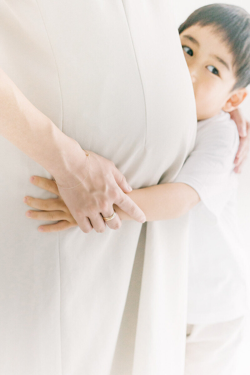 新宿区で撮影。妊婦さんとに長男であるお兄ちゃんが抱き着き、おなかの前で手を添えているおしゃれで可愛らしいマタニティフォト。ホワイトコーデで全身白を基調としたスタイリングで撮影。