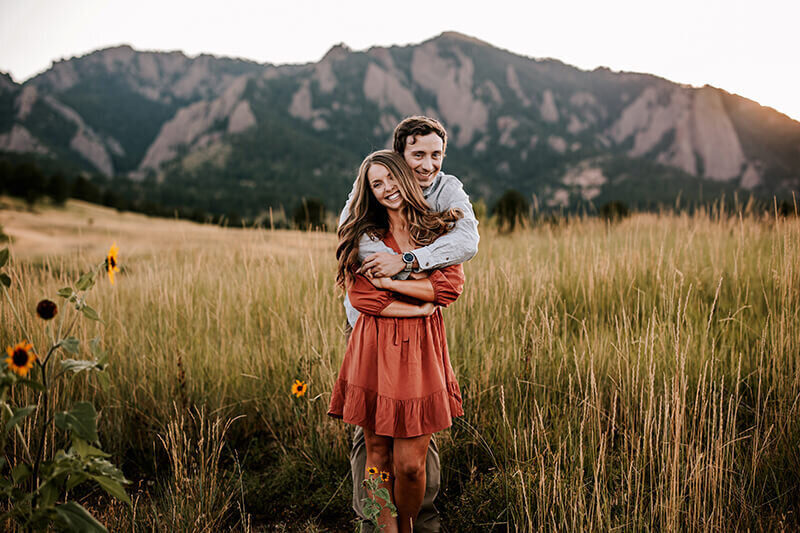 Husband embraces wife from behind in a grassy field with mountains in the background