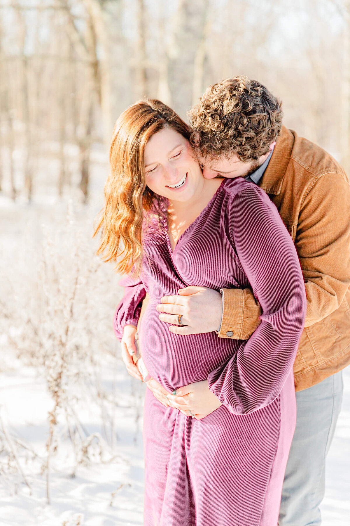 Pregnant woman laughs as her husband holds her baby bump from behind in a snowy park