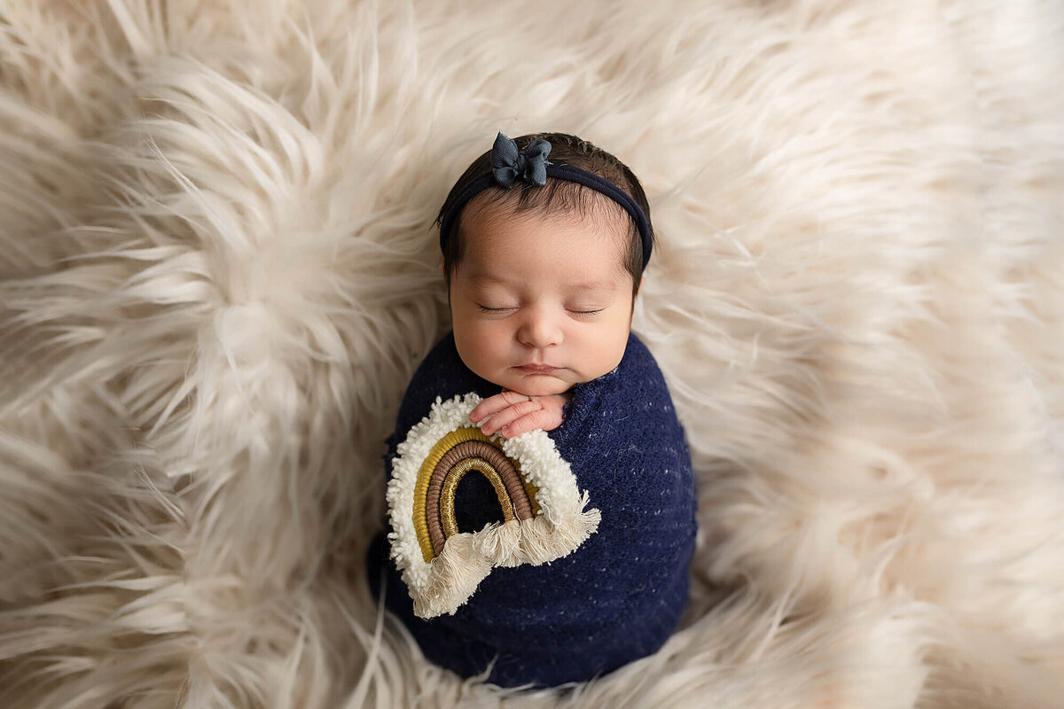 Newborn girl wrapped in blue swaddle holding a golden rainbow.