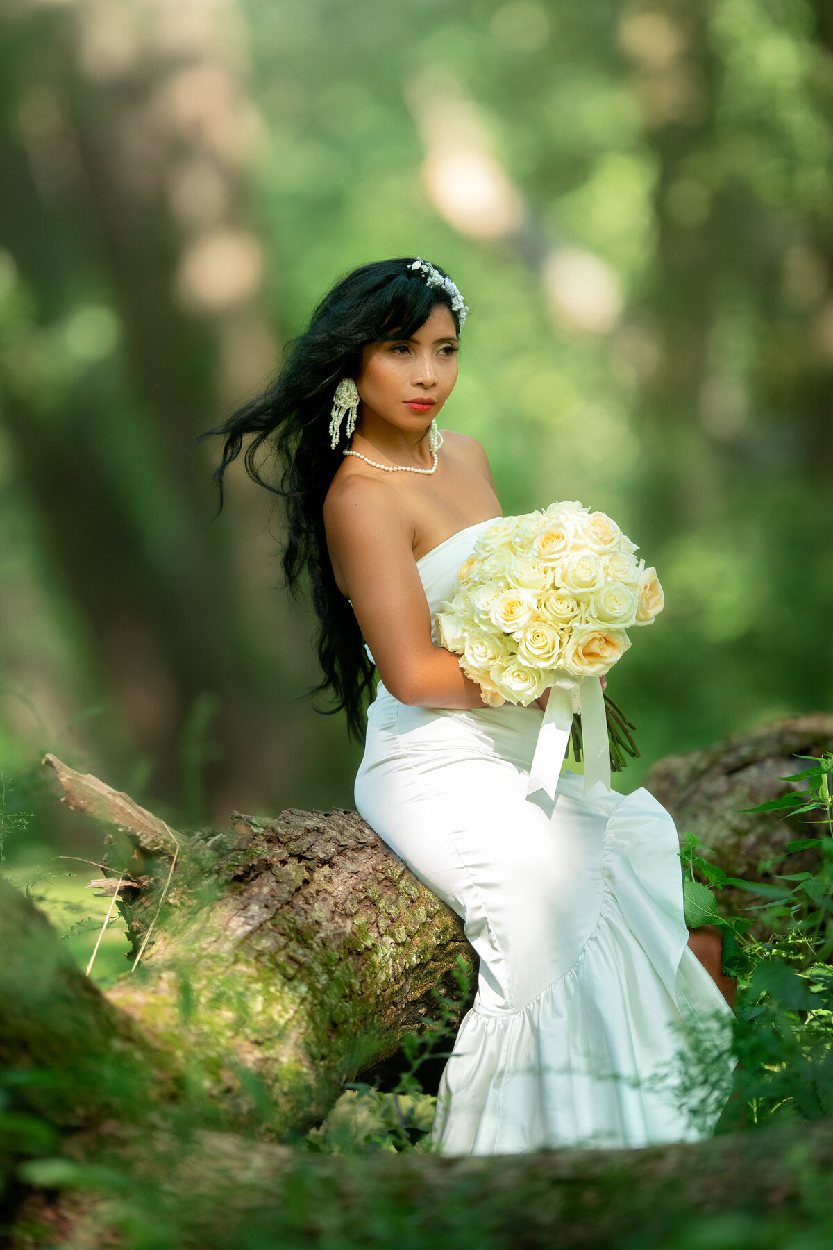 Soon to be wife is posing on the tree log  in a white wedding dress  while holding white roses