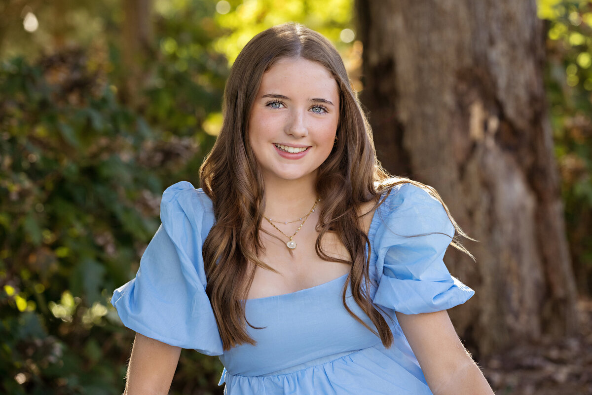 Graceful tween daughter in a flowing blue dress seated on a park bench with soft foliage behind her in South Charlotte NC, relaxed and composed.