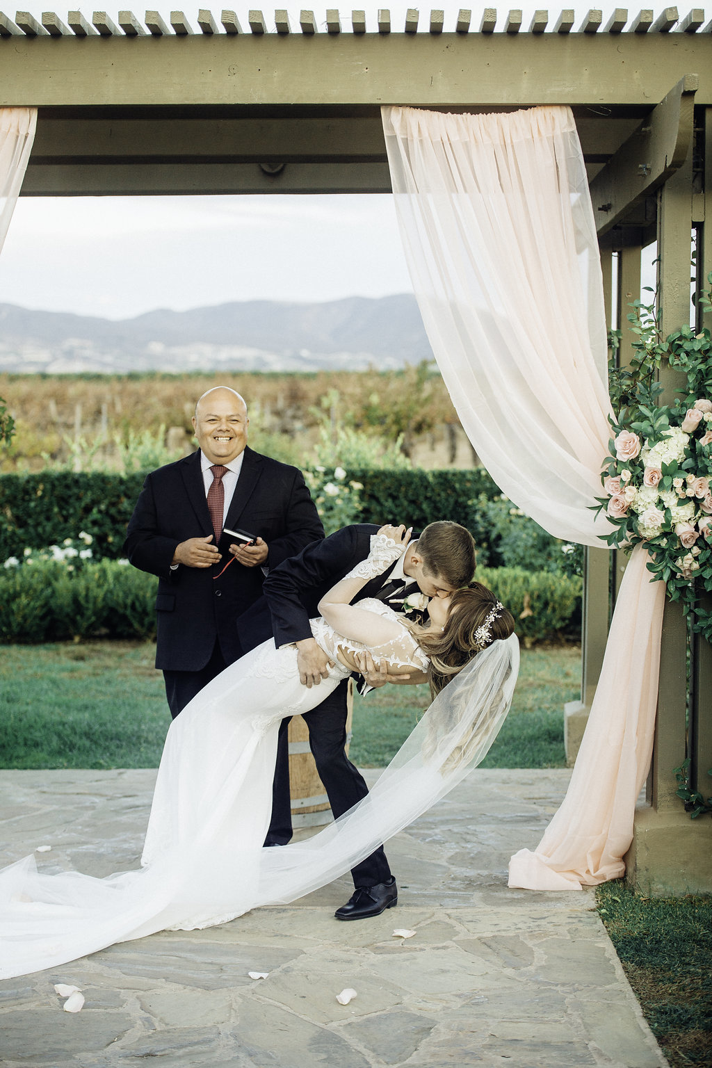 Wedding Photograph Of Pastor Smiling To Bride And Groom Bending While Kissing Los Angeles