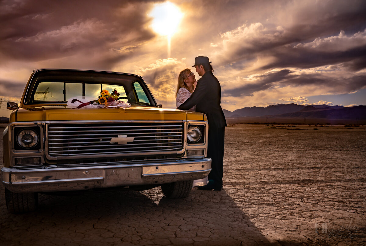 Bride and groom looking lovingly at each other during golden hour with the sun peaking through the fluffly pink and purple clouds with the mountains in the backgroun Dry lake bed elopement bride in lace wedding gown with glasses groom in black suit white dress shirt with  fedora  classic chevy truck gold  restored with golden hour sunlight las vegas wedding photography las vegas elopement las vegas wedding photographers mk delacy photography