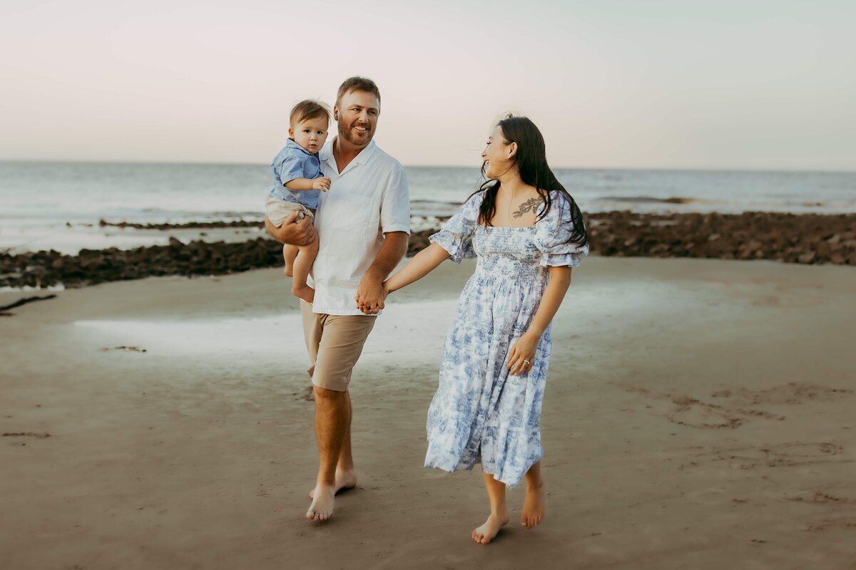 A man and woman walk barefoot on Driftwood Beach, holding hands, with the man carrying a toddler. They appear to be enjoying a calm evening by the sea, perfect for family photography.