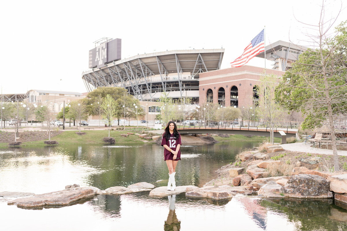 Texas A&M senior girl standing on rocks in maroon jersey and white boots while holding wrist in front of Kyle Field at Aggie Park