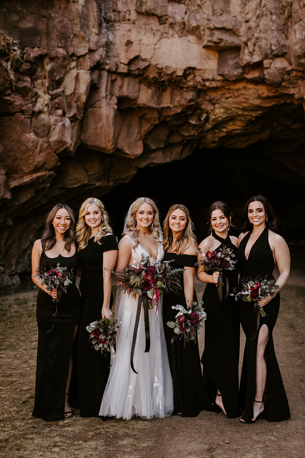 Bride with dark bouquet and black ribbon with bridesmaids in black dresses in front of the caves at Juniper Preserve