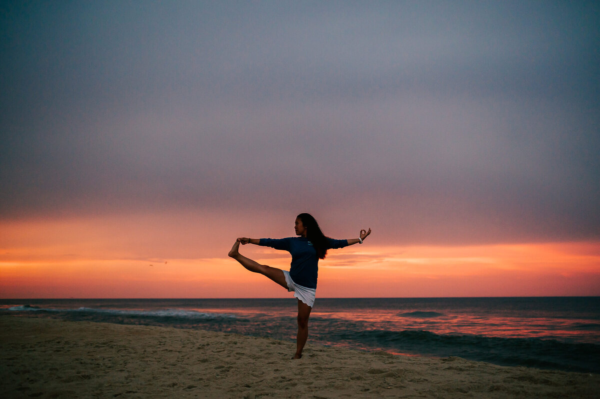 A woman doing toe-holding pose at the beach against a sunset