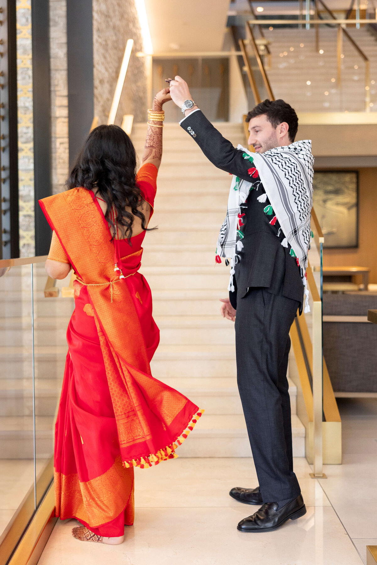 A man and woman dance gracefully by a staircase. The woman is wearing a red and gold saree, and the man is in a suit with a patterned scarf. Theyre holding hands and looking at each other. The setting is an elegant indoor space.