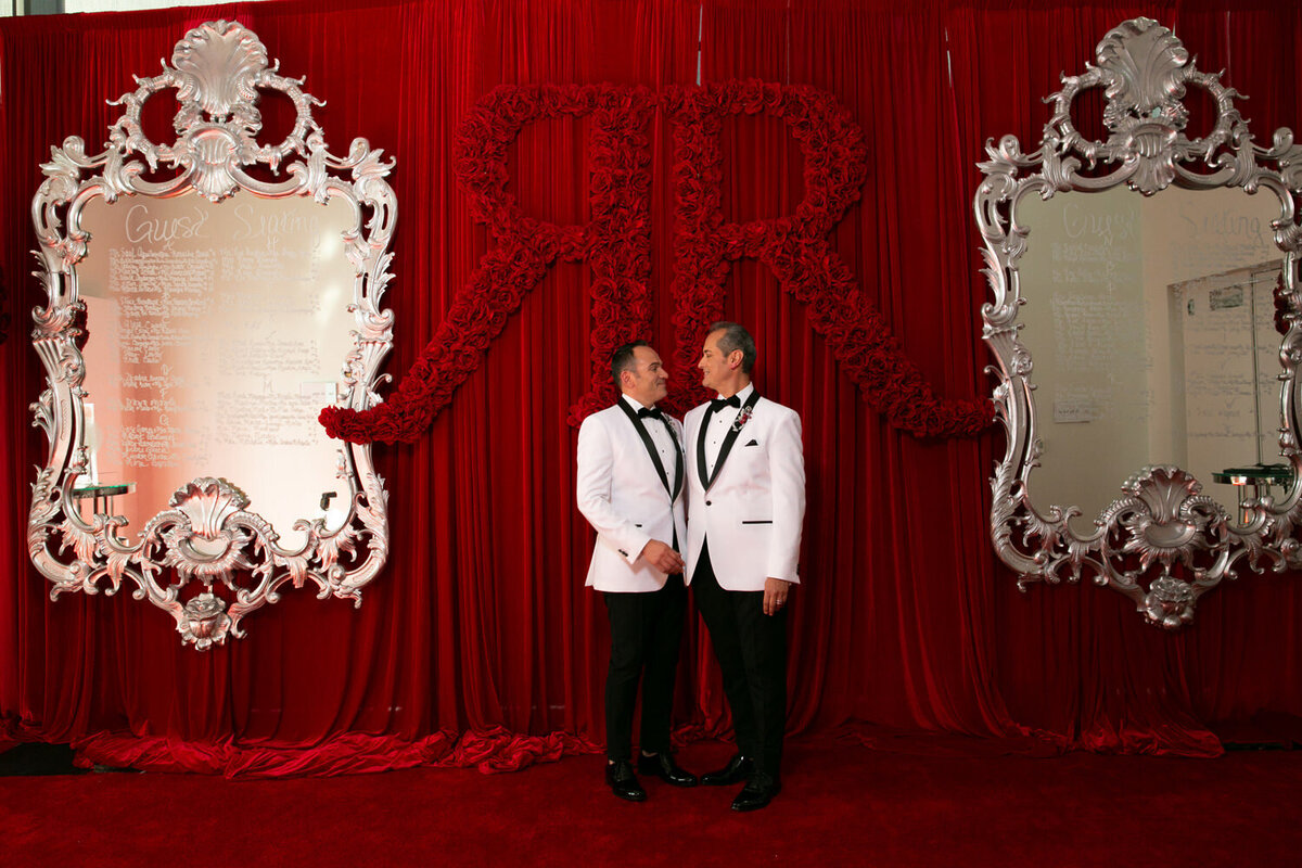 Two grooms standing in front of a wall with two "R"s in red roses behind them