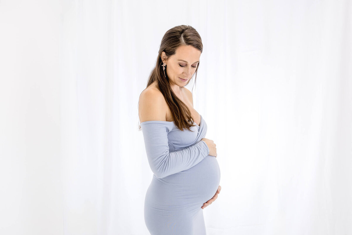 A pregnant woman with long brown hair stands in profile, gently cradling her belly. She is wearing an off-shoulder, form-fitting light blue dress, against a white background, looking down with a serene expression.