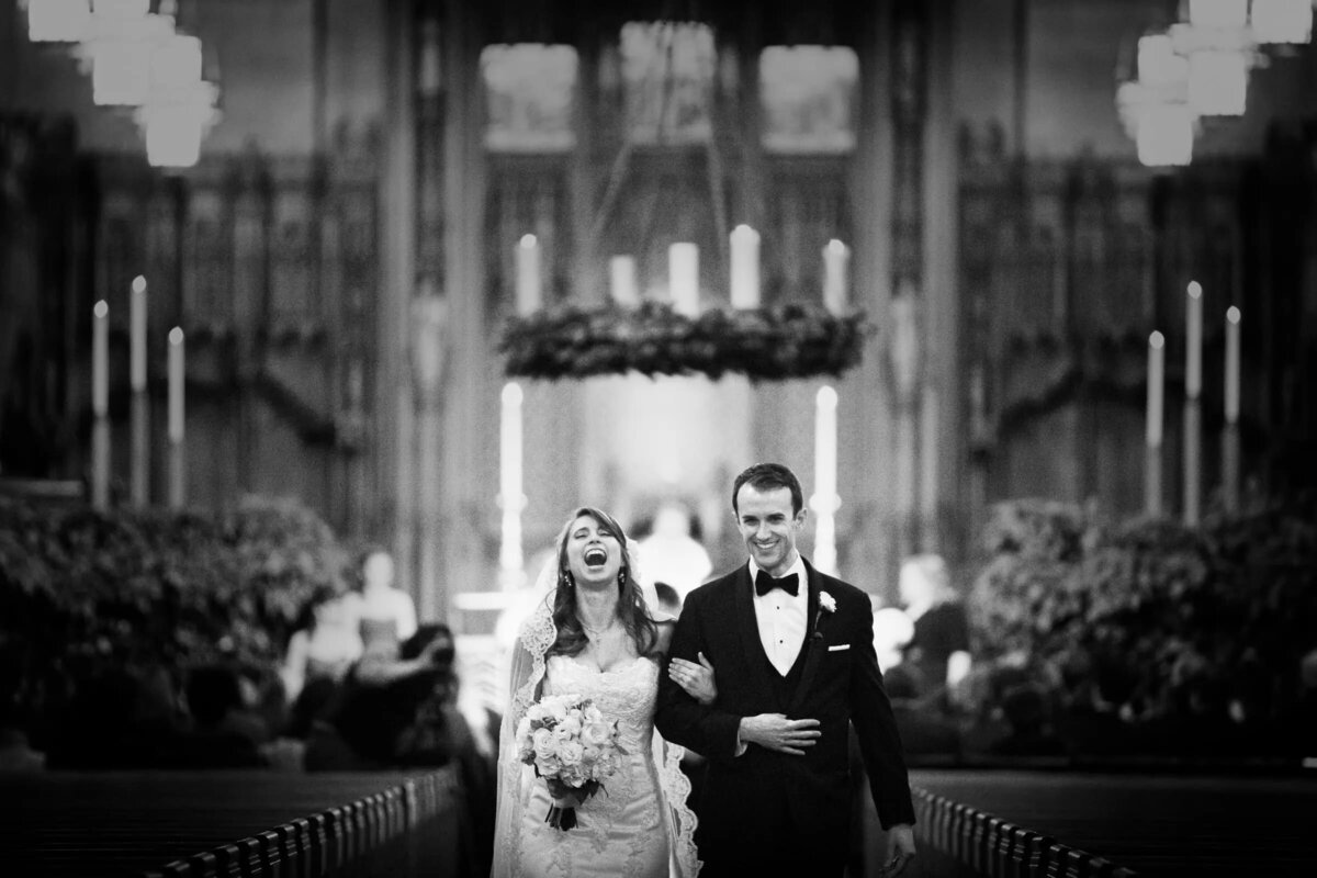 Bride and groom walking joyfully down the aisle during their recessional, in a black and white photo inside a gothic cathedral