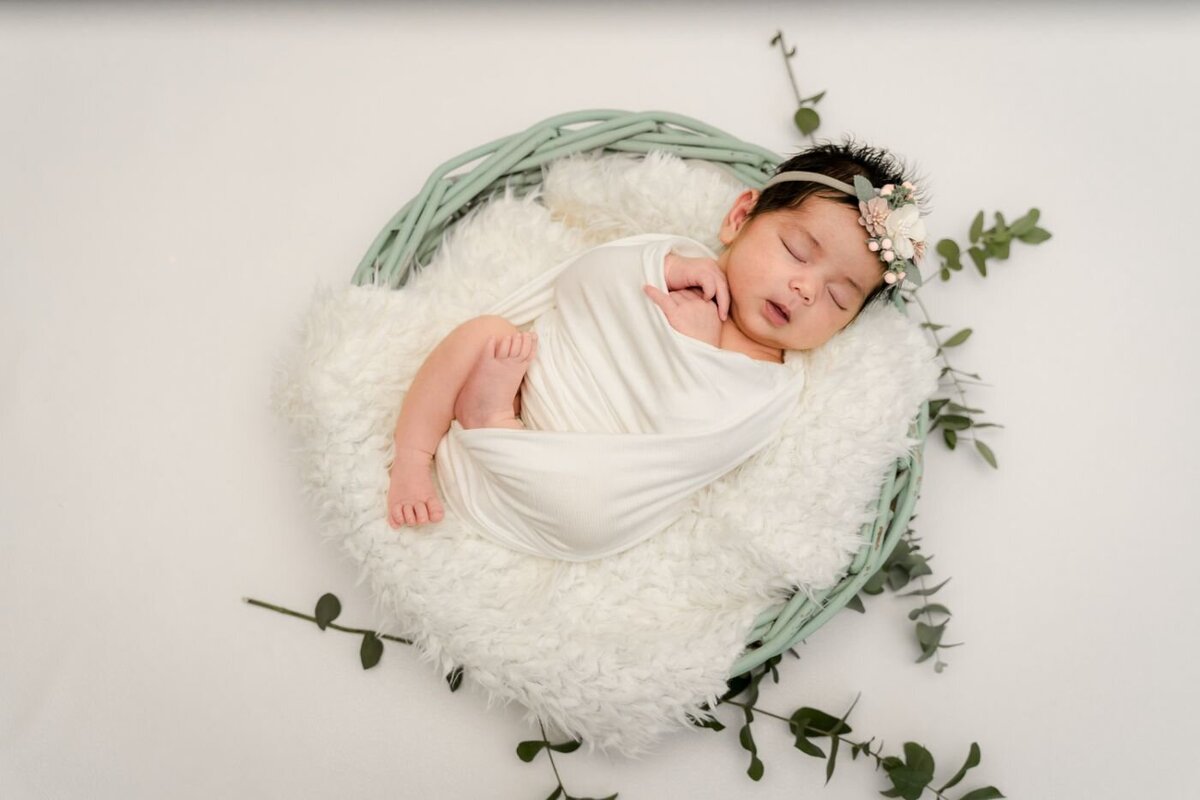 Peaceful newborn wrapped in a white swaddle, sleeping in a mint green basket lined with fluffy white blanket and eucalyptus leaves.
