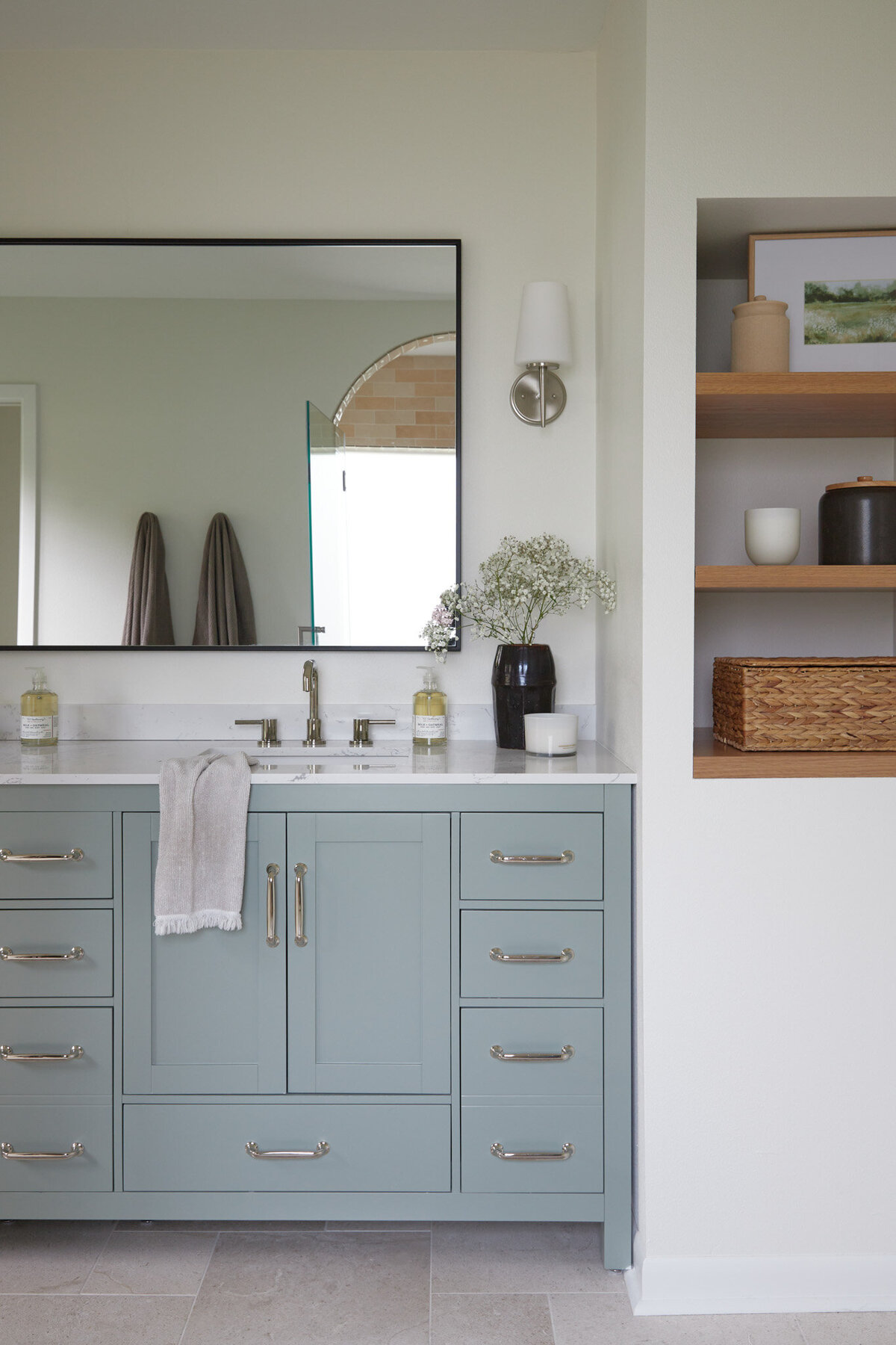 Bathroom with light blue bathroom vanity with silver cabinet pulls. A silver sink faucet with a black mirror above. Next to the sink is a dark brown vase with baby’s breath flowers, a Handsoap pump, and a white candle. Next to the vanity is a white wall with recessed white oak shelves with photos, jars, and a woven basket.