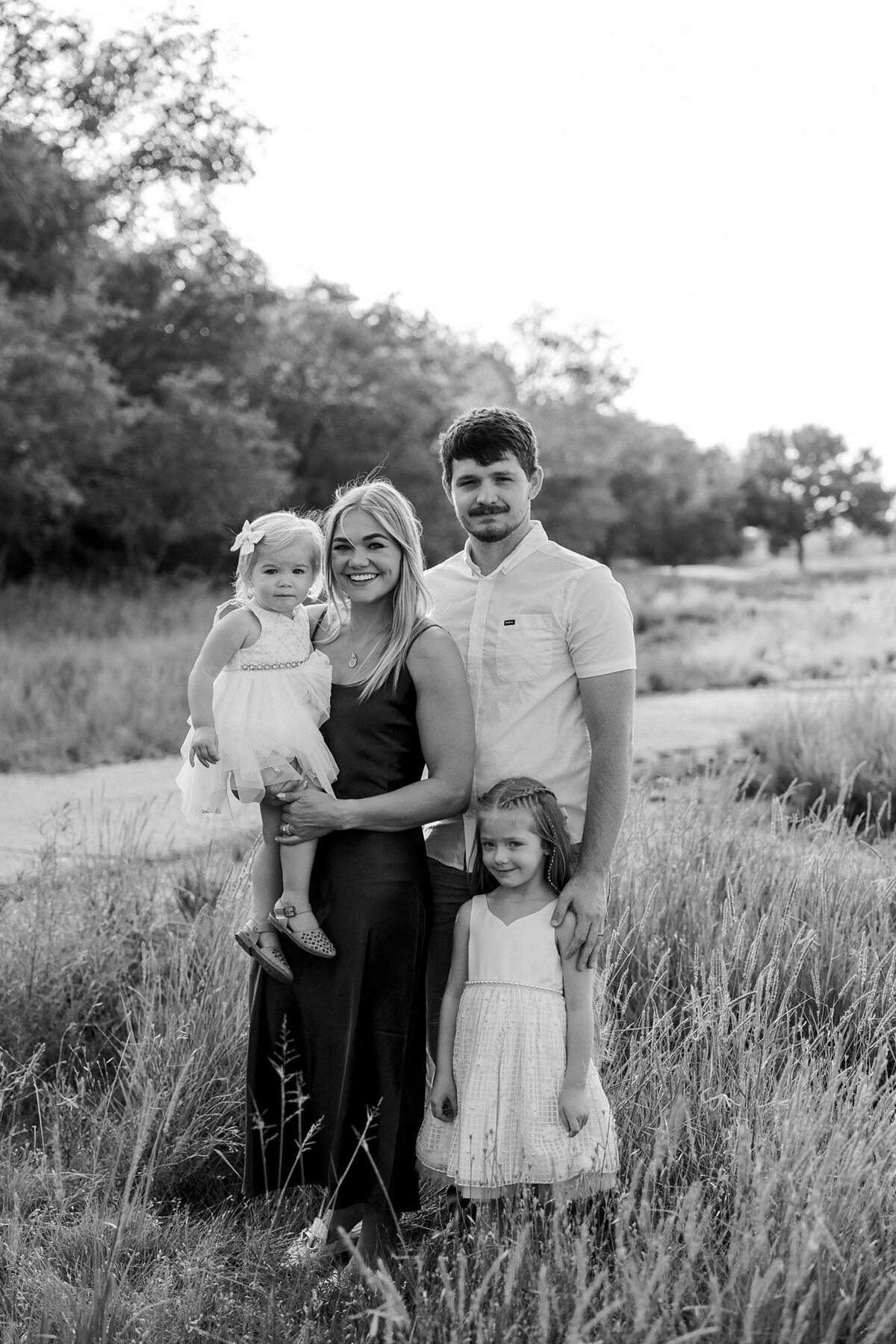 Black and white portrait of a family of four standing in a field in front of a tree row.