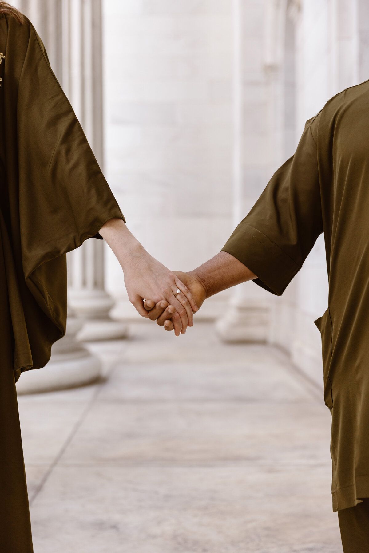 Close-up of an engaged couple gently holding hands in front of tall marble columns, showcasing their rings and matching earth-toned outfits.
