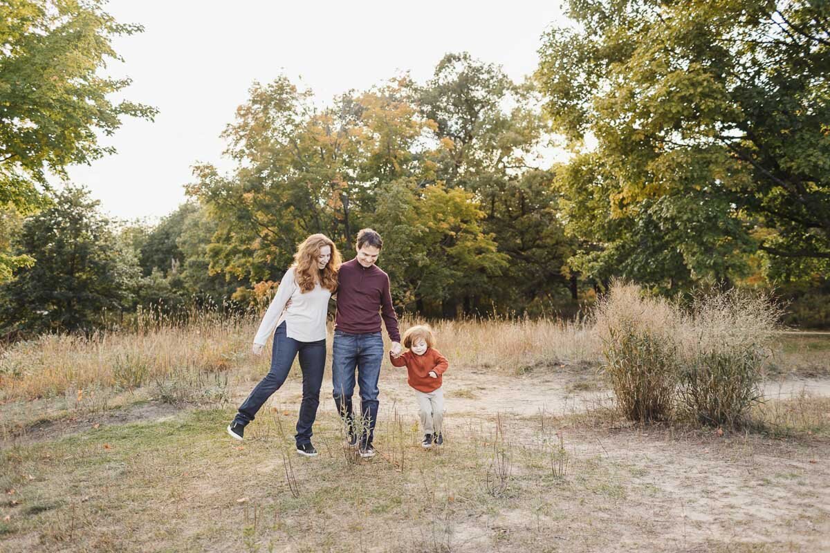 A family of three strolls hand in hand through a grassy, tree-filled park. The child walks between the two adults. It's a sunny day with a clear sky, perfect for capturing memories by a talented Toronto Family Photographer.