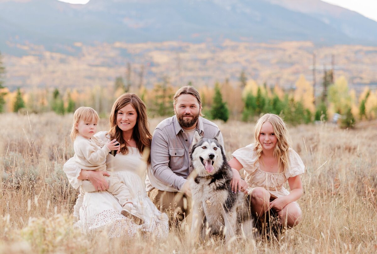 A family of four sit in tall golden grass in a mountain meadow with husky