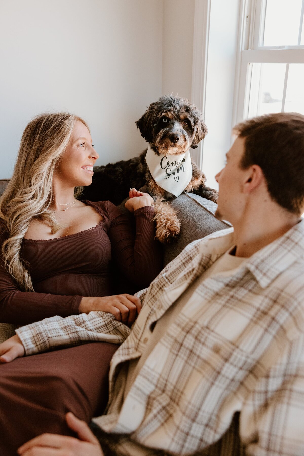 A couple lounges on a couch, smiling at each other while their adorable dog sits between them wearing a white bandana that reads "Coming Soon..."