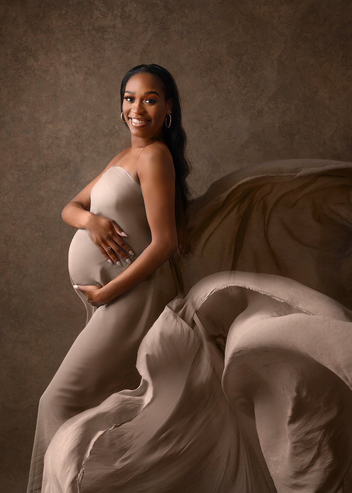 Beautifully posed smiling  pregnant woman, looking at camera, holding her belly, draped in brown silk fabric agains brown textured canvas background in my Phoenix Maternity studio