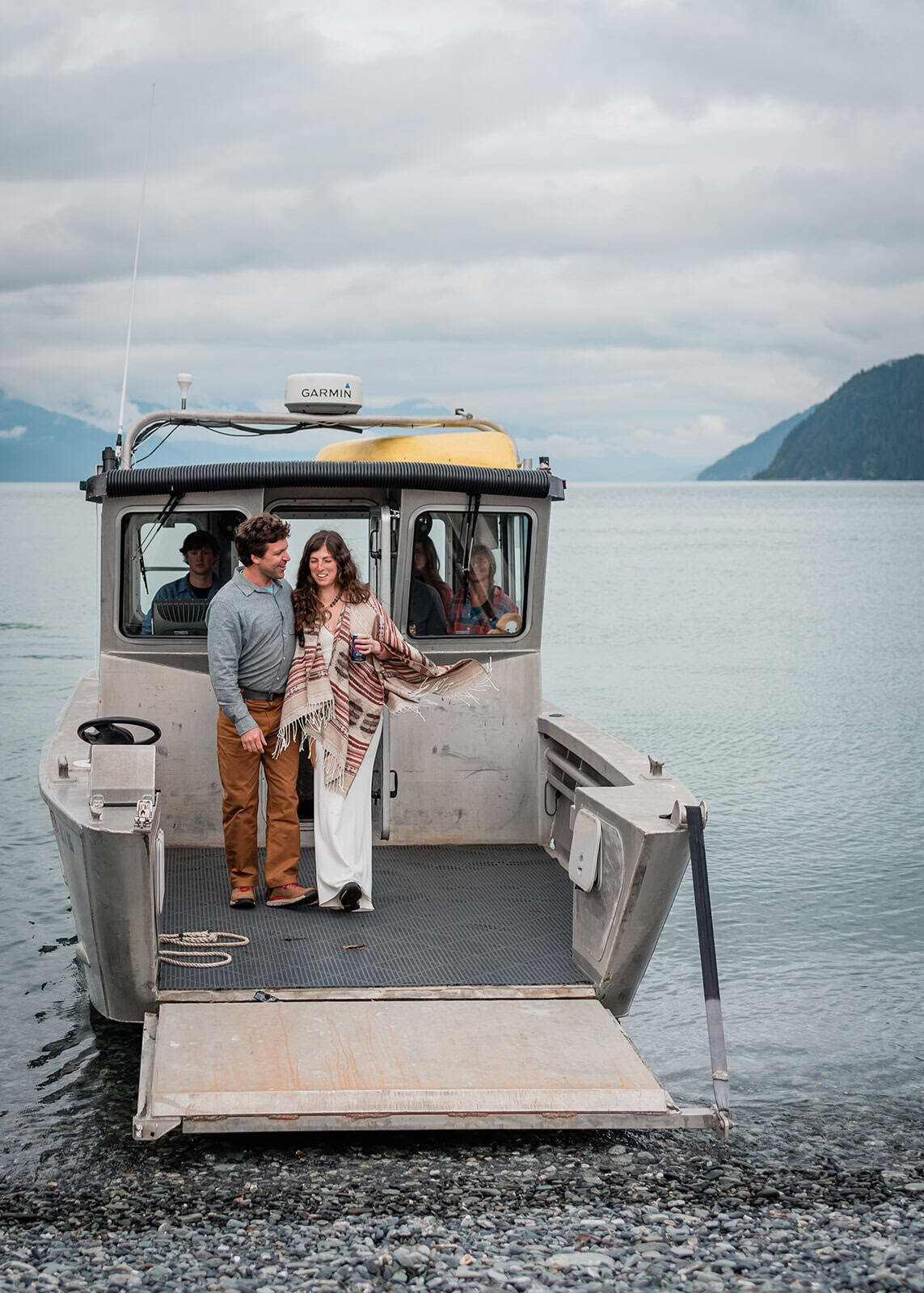 A bride and groom in formal wedding clothes exit off their private water taxi taking them to  the pebble beach and their island vows location during their adventure luxe Kenai Fjords elopement package.  Their guests are standing are slightly visible in the widows of the boat behind them.  Everyone is smiling and laughing. 