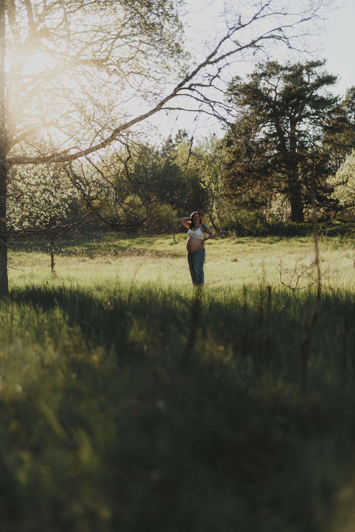En gravid kvinna ståendes i gräset på ett gräsfält i solnedgången lite längre bort under en gravidfotografering i Stockholm