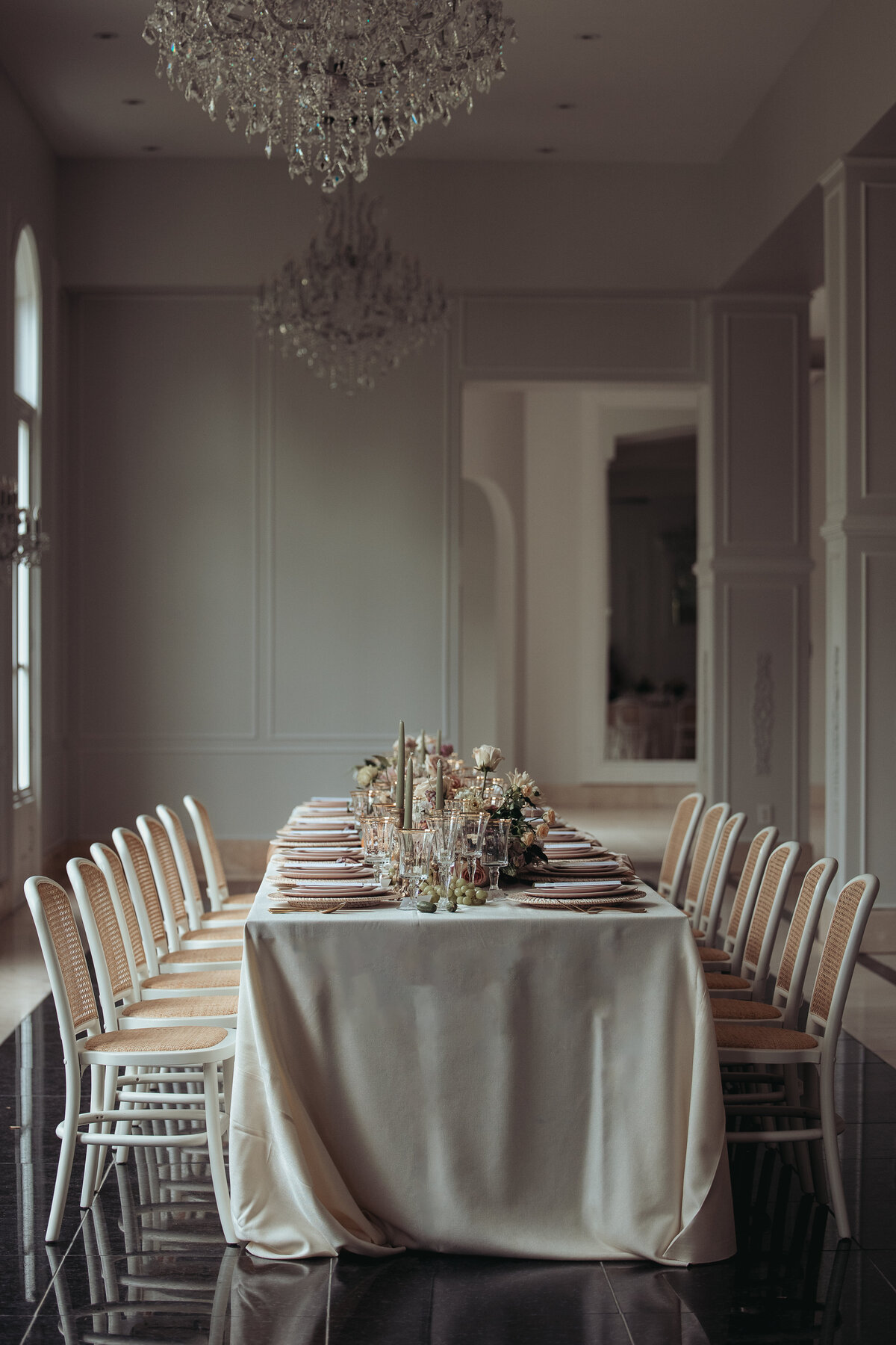 Long reception table with ivory linen, rattan-back chairs, and candlelit centerpiece in an elegant Houston venue.