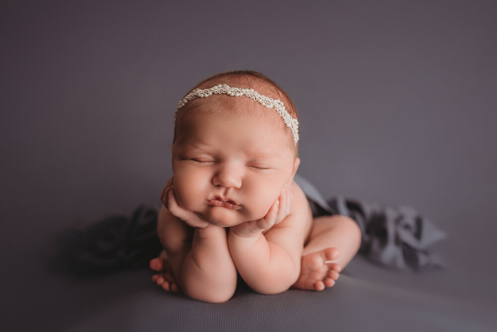 Newborn portrait session with Atlanta, Georgia newborn photographer. 2 week old newborn girl asleep posed on elbows with chin on hands and white pearl headband on blue fabric backdrop.