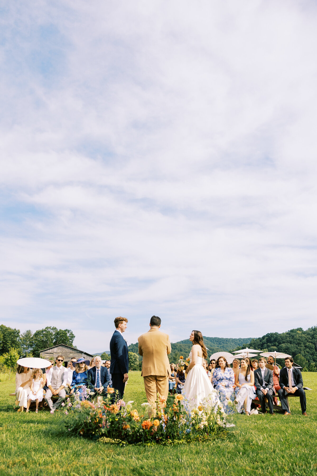 The bride and groom look at each other during their ceremony in front of the happy wedding guests, some using parasols during the summer wedding at Paint Rock Farm a wedding venue in North Carolina, by film photographer Megan Lynn of My Sun and Stars Co.