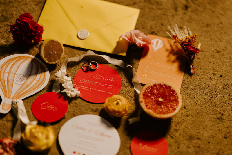 Papeterie de mariage jaune, orange et rouge réhaussée de fleurs et d'agrumes disposée sur un sol ocre par Laura, photographe.