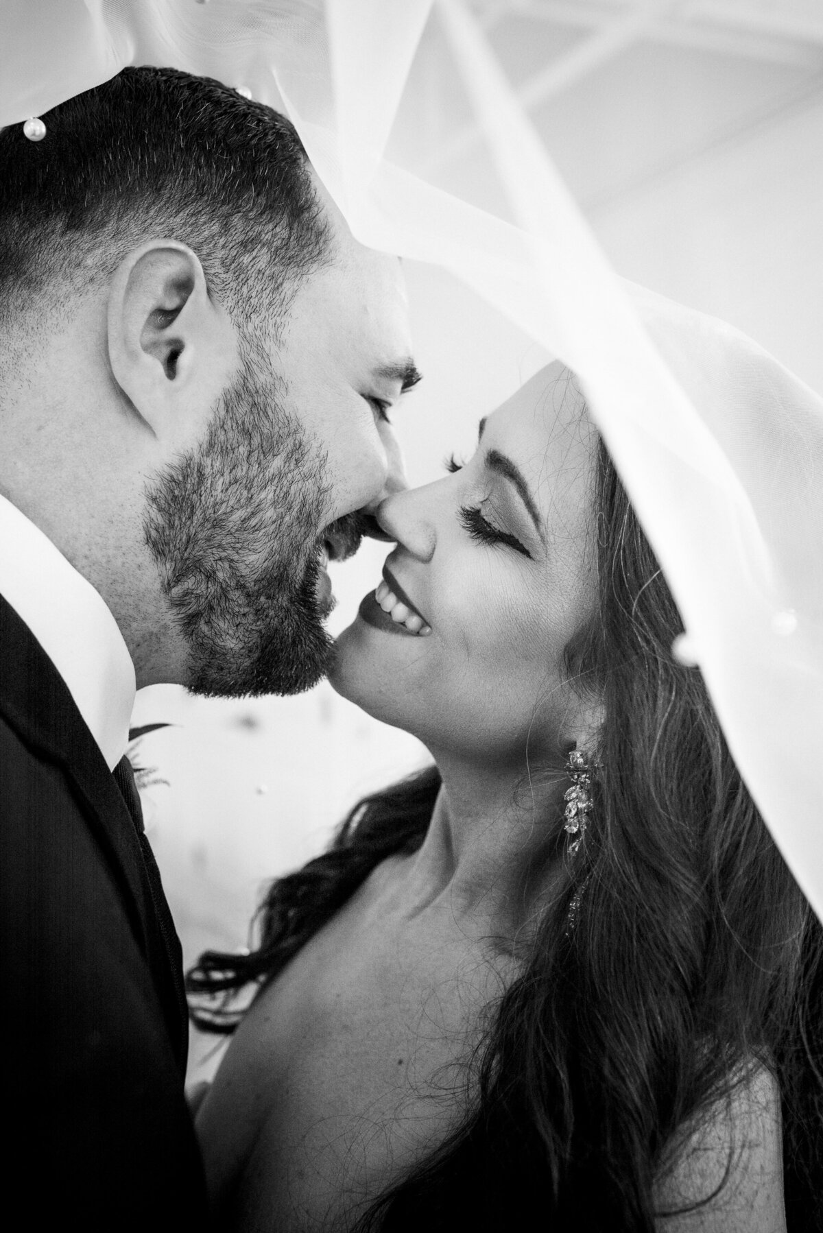 Black-and-white-up-close-vertical-portrait-of-bride-and-groom-smiling-just-before-a-kiss-under-the-bride's-veil-at-Upstairs-Atlanta-by-Charlotte-wedding-photographers-DeLong-Photography
