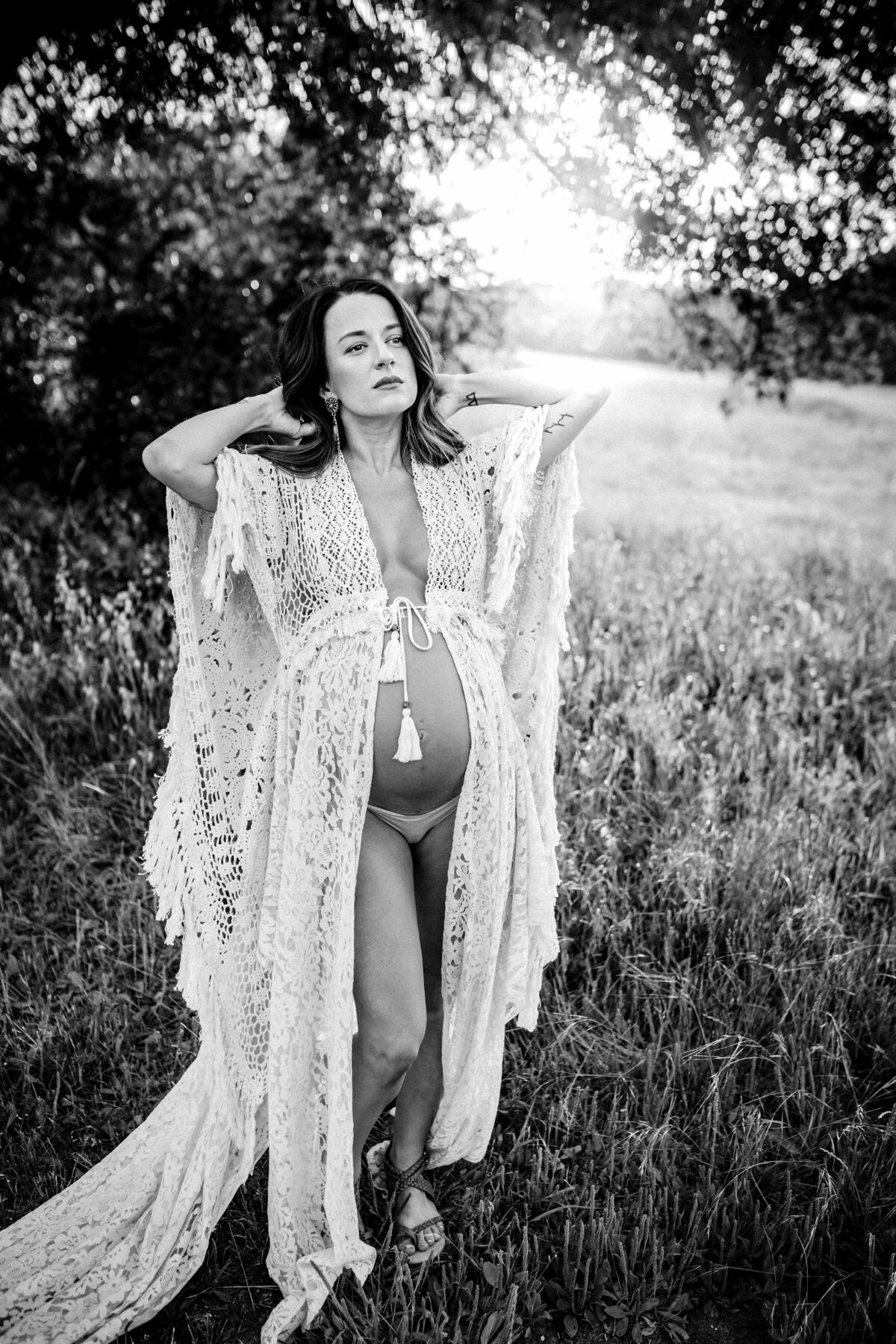 Black and white photo of a pregnant woman standing in a field of grass. She is wearing a macrame robe with her belly exposed while she touches her hair.