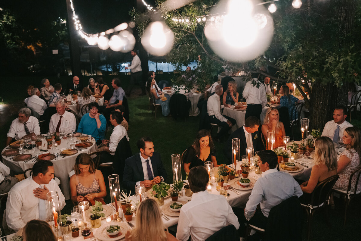A color photograph of the outdoor dinner reception during Colby and Jack’s wedding at Commodore Perry Estate in Austin, Texas. The view is from slightly above, showing the guests seated at their tables in conversation. The tables are surrounded by trees and cafe lights overhead light the scene. Wedding photography taken by Stacie McChesney/Vitae Weddings.