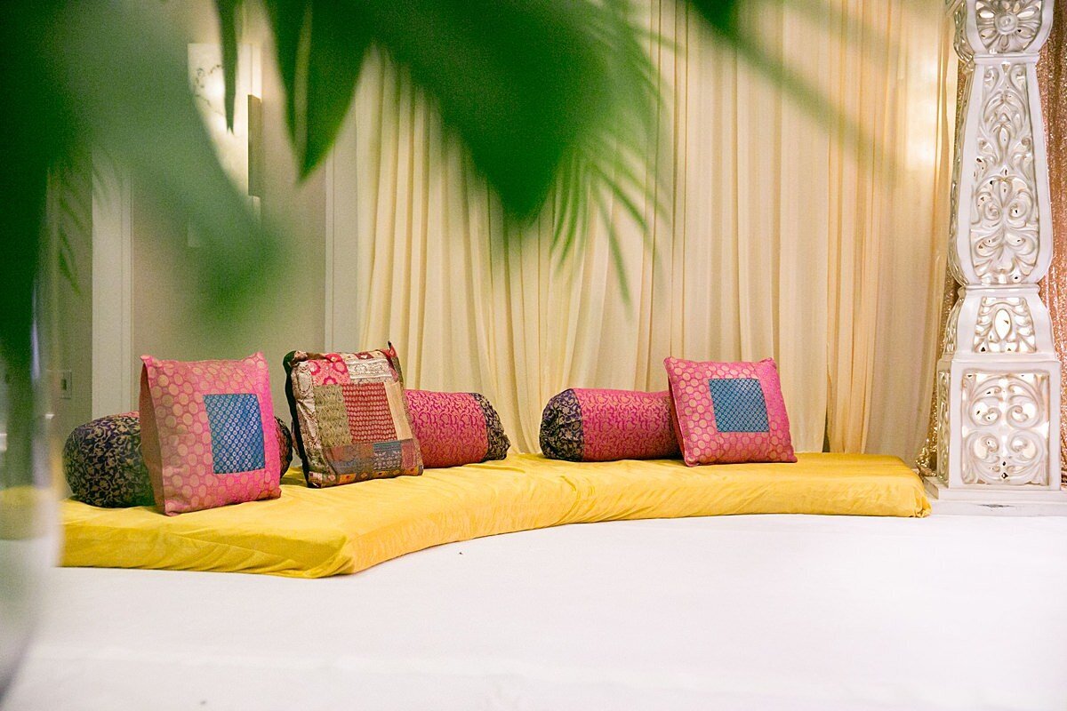 Magenta, orange, blue and gold decorative pillows sitting on a long yellow cushion underneath an ivory mandap with gold filagree detail and large green palm fronds.