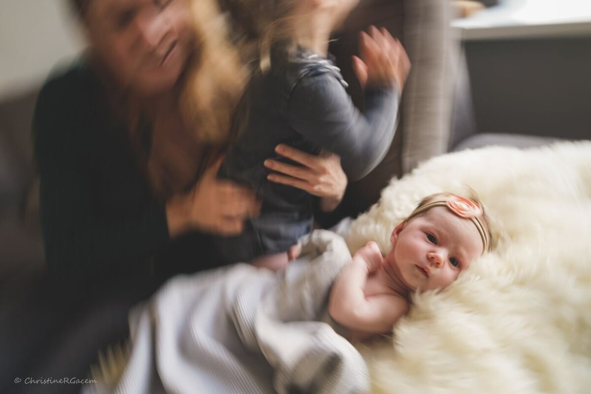 Newborn with family in home session – A wide-eyed newborn lies on a fluffy white blanket while older sibling moves playfully and mother reaches in, creating a candid, motion-blurred moment indoors.