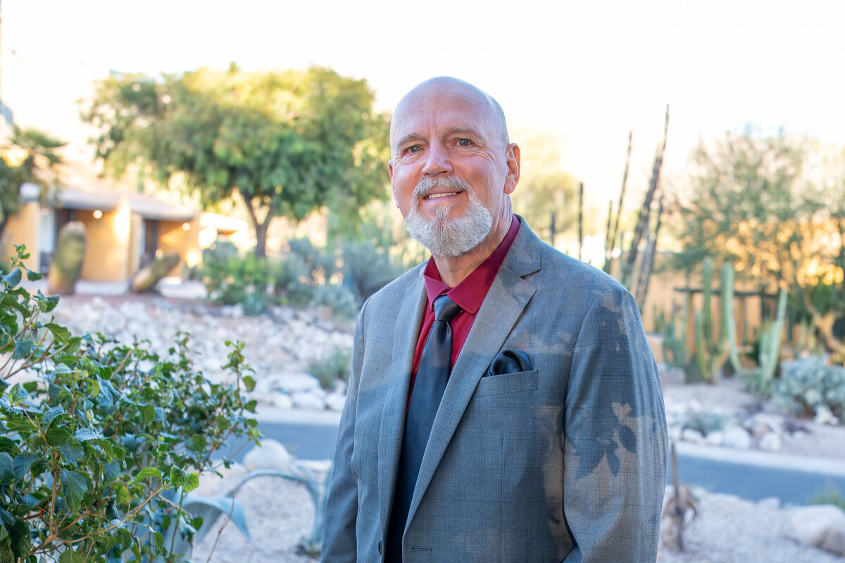 Man in a gray suit and red shirt smiling outdoors in a desert garden setting, photographed by Vyrl Photo for a professional Tucson business portrait.