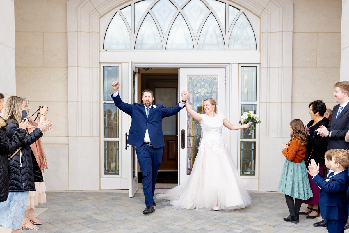 Bride and groom hold hands and raise their arms triumphantly while exiting the Taylorsville Utah LDS Temple doors, surrounded by cheering family and friends.