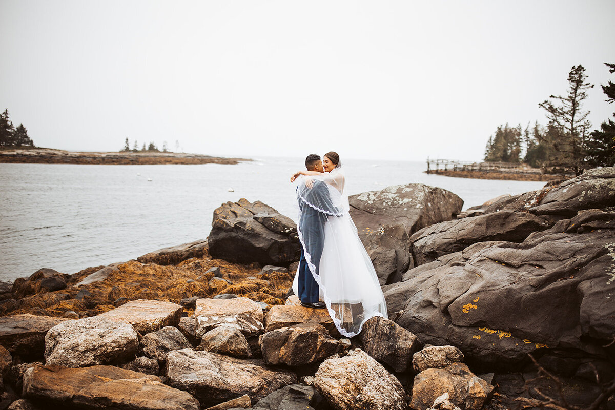 Bride and groom embracing on the rocky shoreline with ocean views during their Maine coastal wedding.
