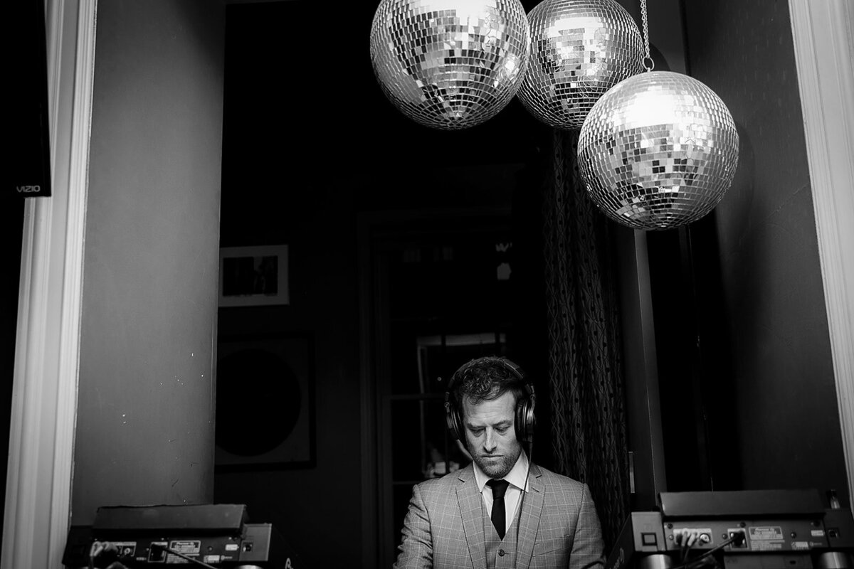 Black-and-white photo of a DJ in a suit performing under disco balls.