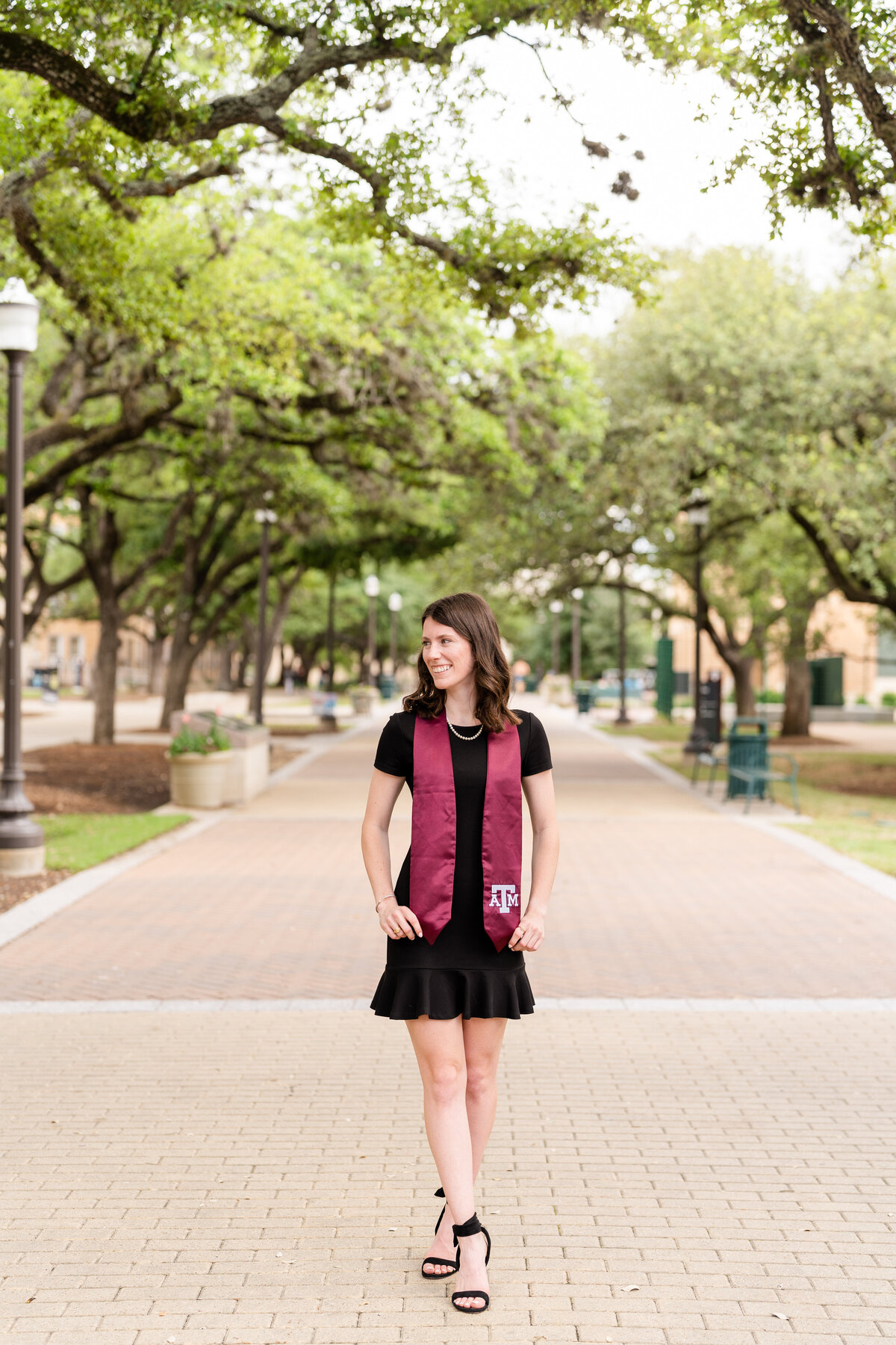 Texas A&M senior girl wearing little black dress and holding Aggie stole while smiling away under the trees of Military Plaza