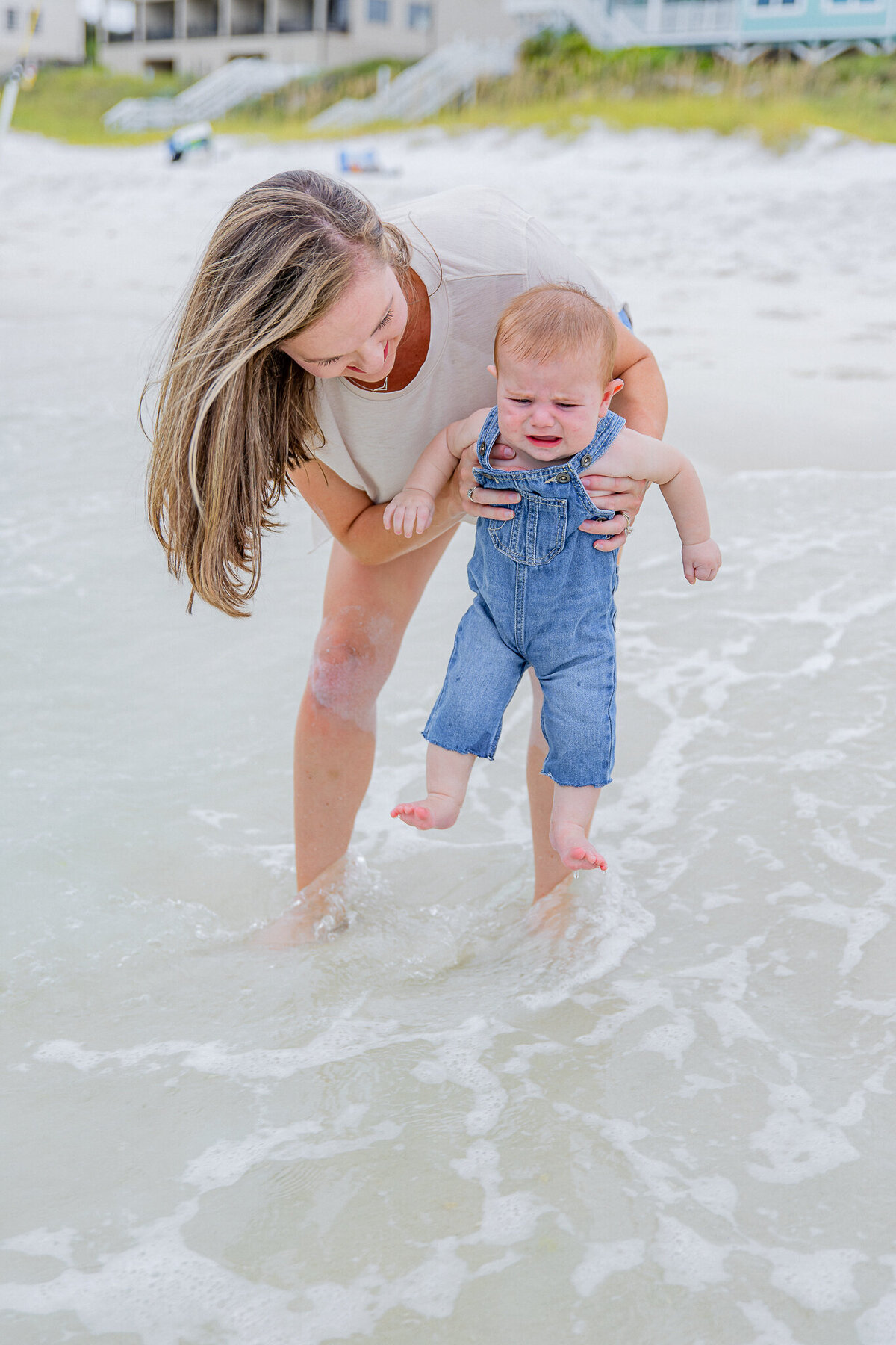 A joyful mother and her two young children play in the waves at Orange Beach, AL. The mother holds her baby securely while her curly-haired daughter, wearing a pink bow, laughs as the foamy ocean water swirls around their feet. Beachfront vacation homes provide a serene coastal backdrop in this timeless black-and-white family portrait