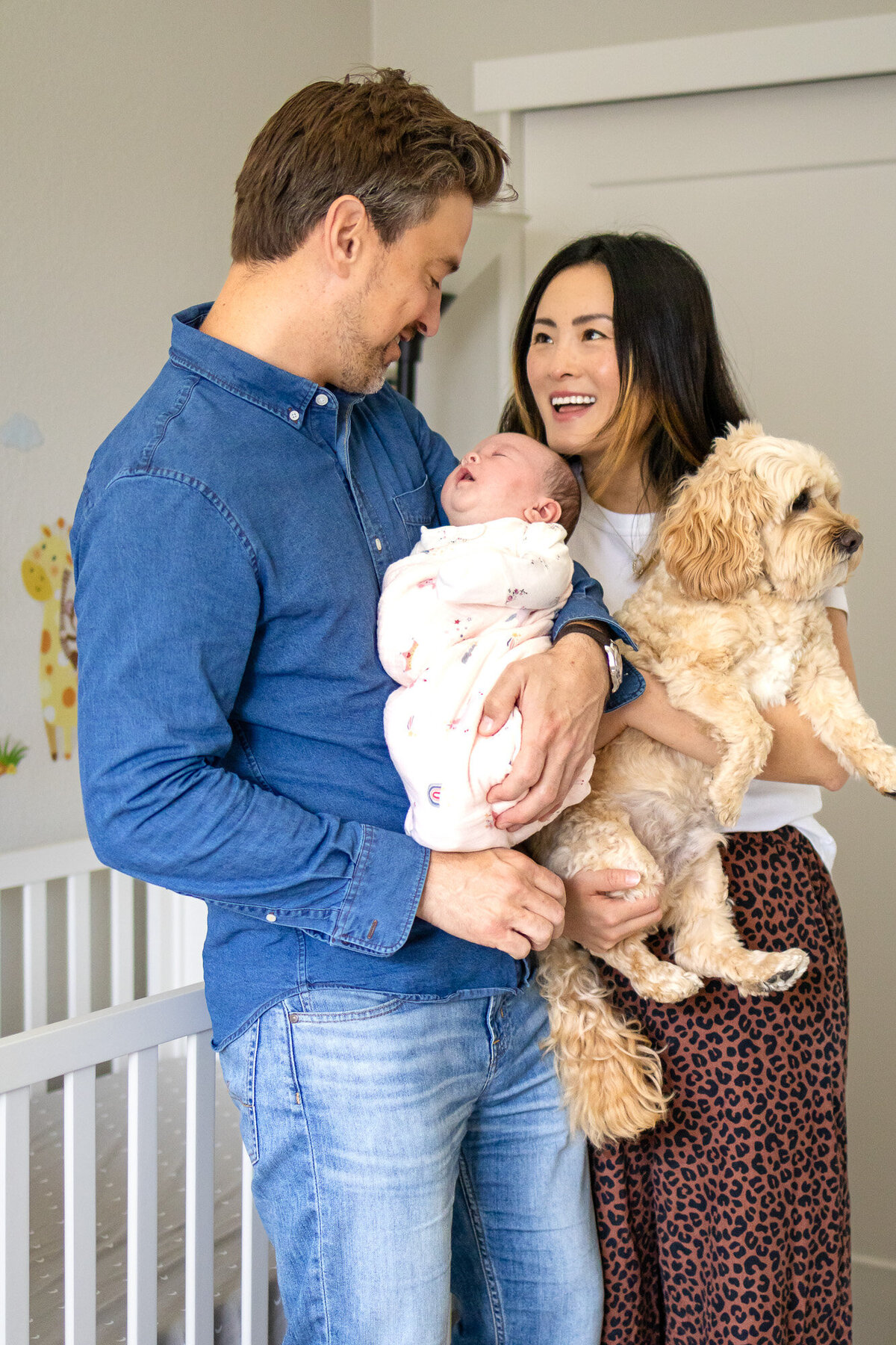 Father cradling newborn while mother holds family dog, smiling together in nursery – by San Francisco newborn photographer.