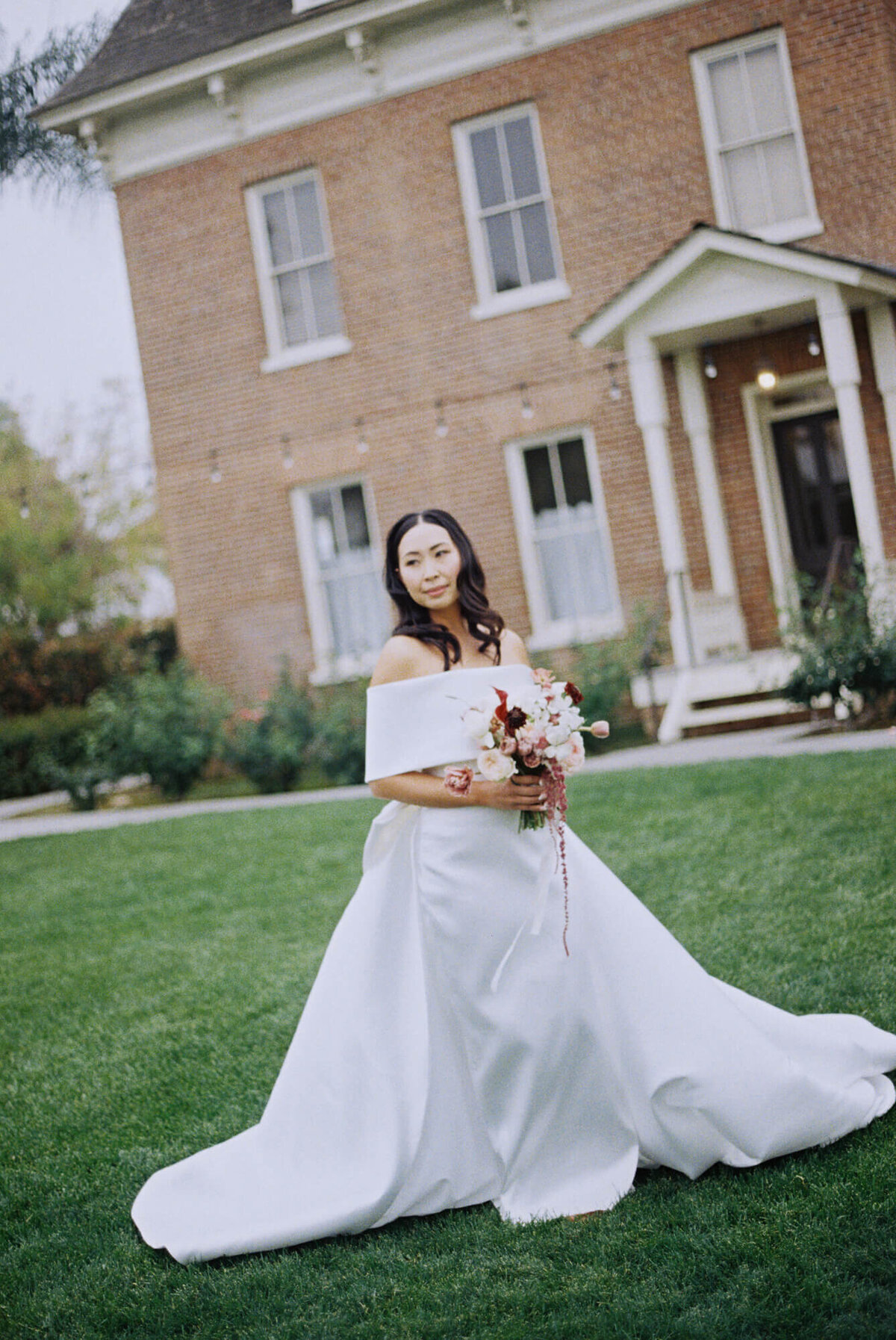 Bride in an elegant white off-the-shoulder gown holds a pink bouquet, standing on green grass in front of a brick house