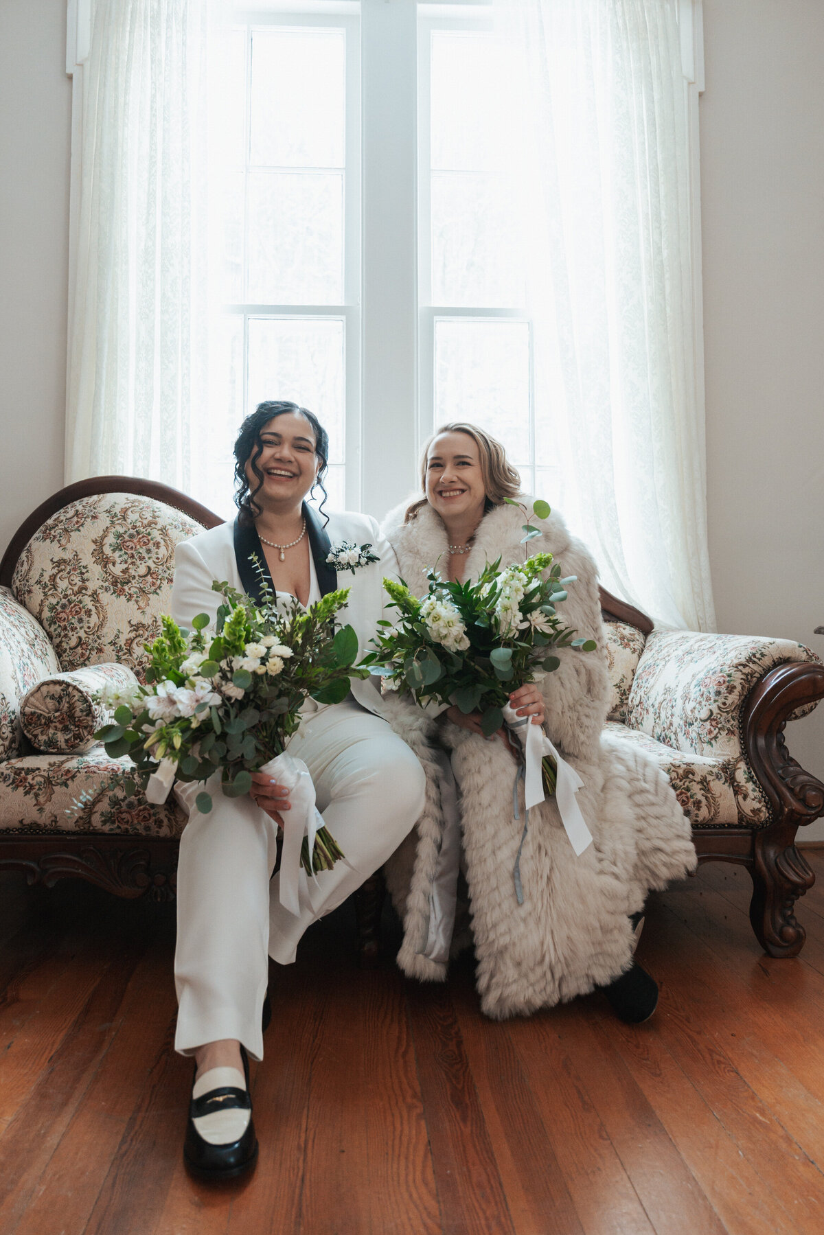 Two women sit on an antique sofa with their large bouquets in their laps 