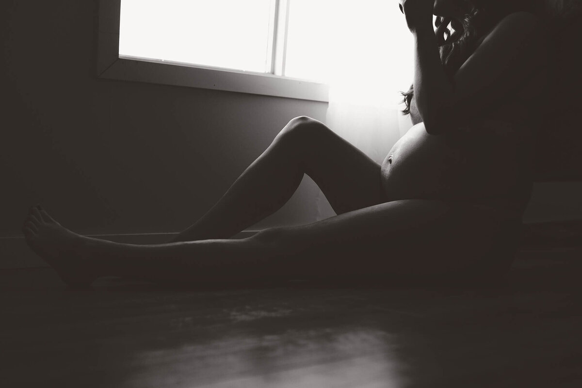 A darker maternity portrait. A pregnant woman in her underwear sits on her bedroom floor with one leg flat and one knee up. A bright window is behind her. Captured by Brittany Danielle Photography.