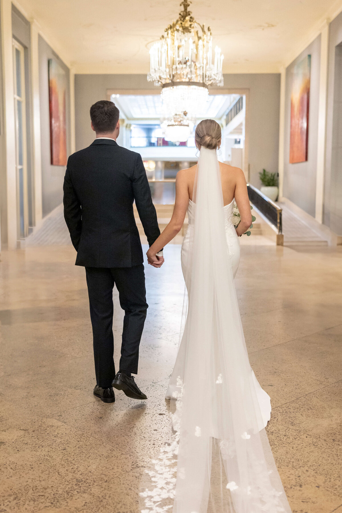 A couple is walking away, hand in hand, in an elegantly decorated hallway. The bride is wearing a long white gown with a veil, and the groom is in a dark suit. Chandeliers hang from the ceiling, and abstract art adorns the walls.