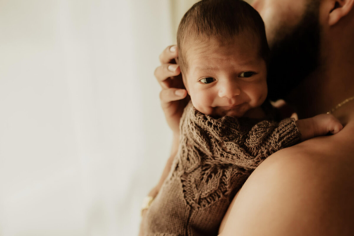 Baby boy resting on his fathers chest wearing a brown knit romper.