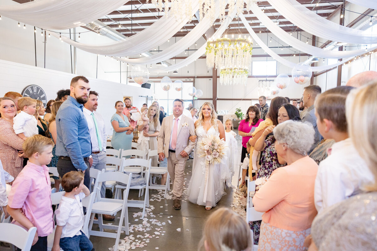 The bride walks down the aisle with her father, surrounded by family and friends, under a beautifully decorated ceiling draped in white fabric and twinkling lights. The venue, Maplewood Events in Spanish Fork, Utah, provides a rustic yet elegant atmosphere for this unforgettable moment. Captured by Cassie Lee Photography.
